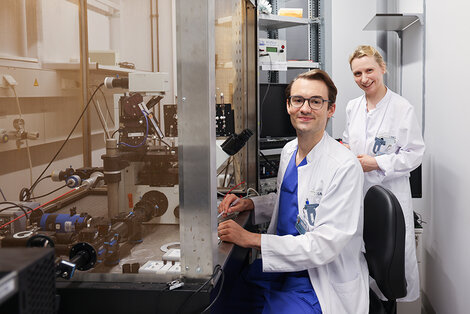 Two scientists in lab coats work in a laboratory; one operates equipment at a workstation while the other stands nearby, both looking at the camera.