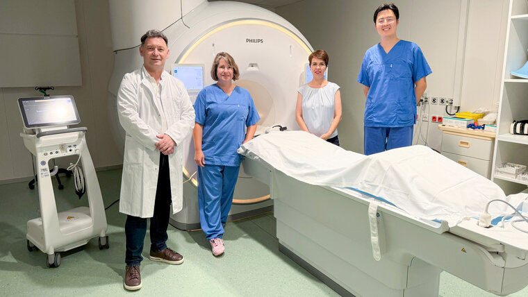 Four medical professionals stand in front of an MRI machine in a hospital room. 