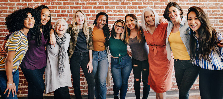 A group of ten women of different ages and backgrounds stand smiling arm in arm in front of a brick wall. The women appear friendly and positive, wearing casual clothes and expressing a strong bond and community.