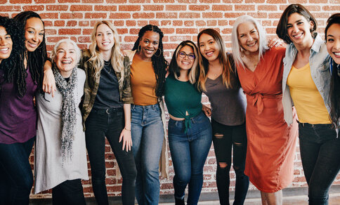 A group of ten women of different ages and backgrounds stand smiling arm in arm in front of a brick wall. The women appear friendly and positive, wearing casual clothes and expressing a strong bond and community.
