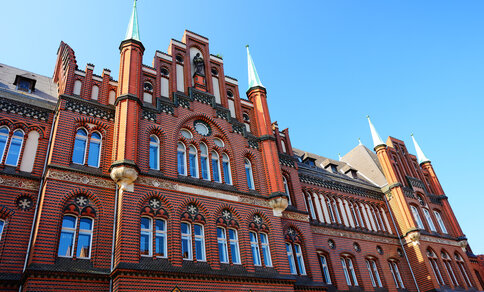 red brick building with intricate patterns, arched windows, and green spires under a clear blue sky.