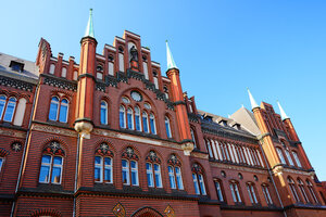 red brick building with intricate patterns, arched windows, and green spires under a clear blue sky.