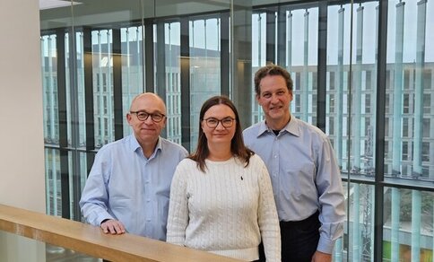 Drei Personen in einem modernen Gebäude mit Glasfassade: links ein Mann(Prof. Dr. med. Christoph Knosalla), eine Frau(Dr. Mariya Kucherenko ) in der Mitte und ein Mann rechts( Prof. Dr. med. Wolfgang Kübler), alle in hellblauer Kleidung und weißem Pullover.