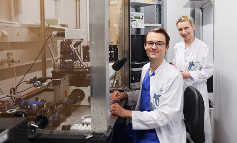 Two scientists in lab coats work in a laboratory; one operates equipment at a workstation while the other stands nearby, both looking at the camera.