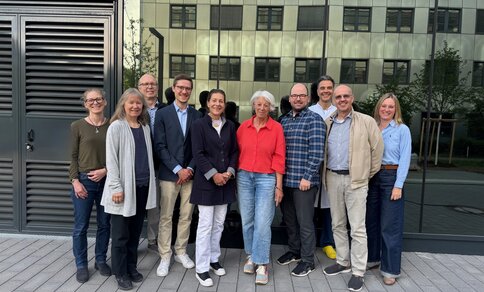 Eine Gruppe von 10 Personen steht lächelnd vor einem Gebäude. Die Stimmung ist freundlich.