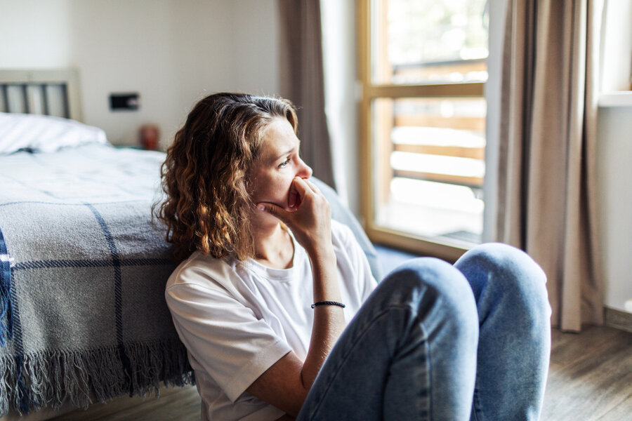 A woman sits deafly on the floor in a brightly lit room near a window. She is wearing a white T-shirt and jeans, her arms resting around her knees as she gazes expressively into the distance.