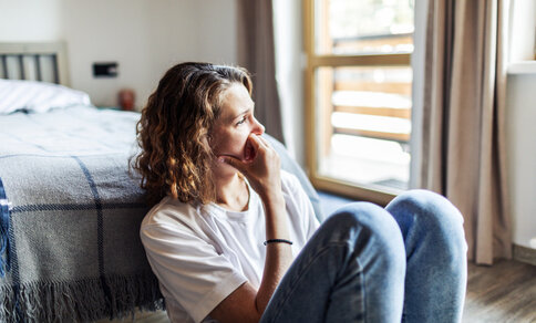 A woman sits deafly on the floor in a brightly lit room near a window. She is wearing a white T-shirt and jeans, her arms resting around her knees as she gazes expressively into the distance.