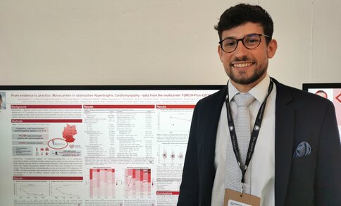 Photo of Dr Jan Kölemen standing in front of his scientific poster at the ESC Congress, titled "From evidence to practice: Mavacamten in obstructive Hypertrophic Cardiomyopathy – data from the multicenter TORCH-Plus-DZHK21". He is wearing glasses, a dark suit, light blue tie, and an ESC Congress lanyard with name badge, smiling at the camera.