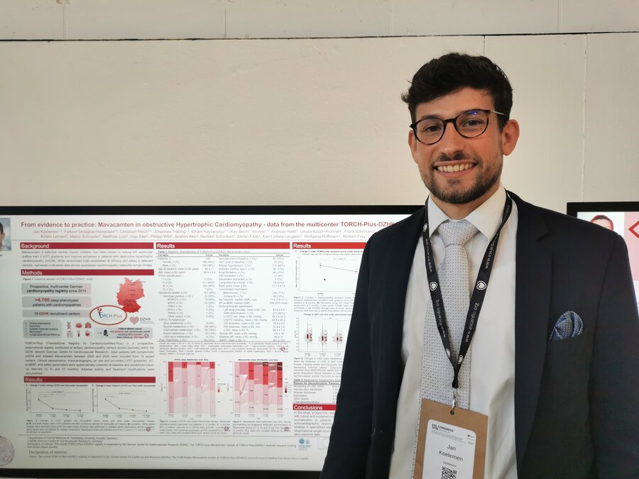 Photo of Dr Jan Kölemen standing in front of his scientific poster at the ESC Congress, titled "From evidence to practice: Mavacamten in obstructive Hypertrophic Cardiomyopathy – data from the multicenter TORCH-Plus-DZHK21". He is wearing glasses, a dark suit, light blue tie, and an ESC Congress lanyard with name badge, smiling at the camera.