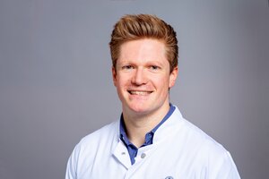 Smiling person in a white lab coat with a blue shirt underneath, set against a neutral gray background.