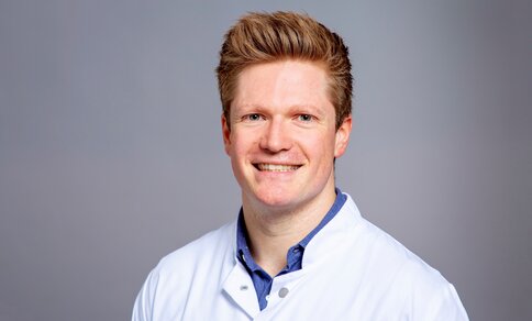 Smiling person in a white lab coat with a blue shirt underneath, set against a neutral gray background.