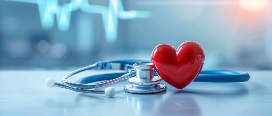The image shows a red heart model next to a stethoscope on a light-coloured, blurred surface. Blurred ECG lines can be seen in the background, indicating a medical environment. It symbolises the connection between heart health and medical diagnostics.