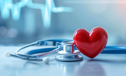 The image shows a red heart model next to a stethoscope on a light-coloured, blurred surface. Blurred ECG lines can be seen in the background, indicating a medical environment. It symbolises the connection between heart health and medical diagnostics.