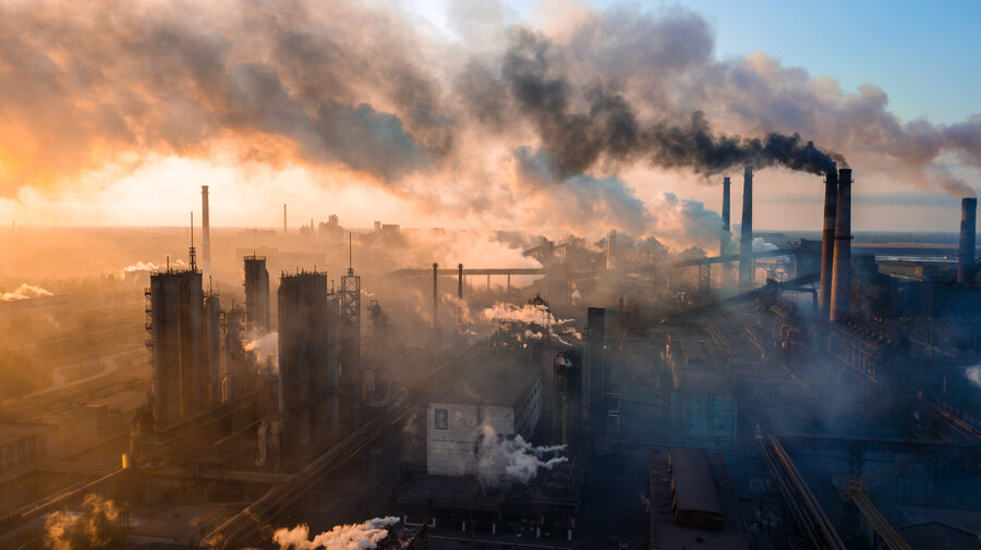 Aerial view of an industrial area at sunset, with several factories emitting thick, dark smoke. 