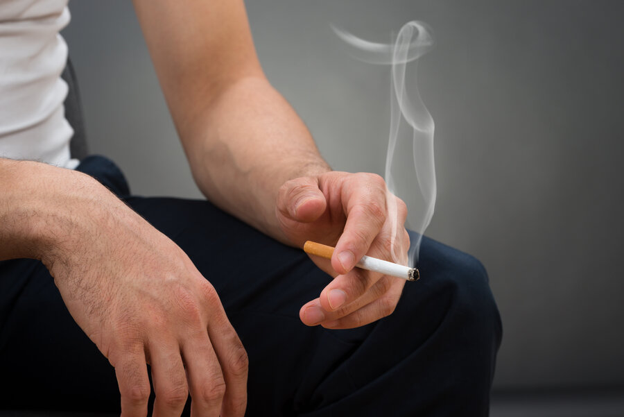 Close-up of a seated person with their hands resting on their knees, holding a lit cigarette between their fingers, with smoke visibly rising.