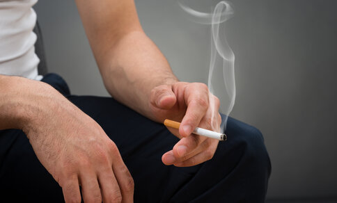 Nahaufnahme einer sitzenden Person mit den Händen auf den eigenen Knien, die eine brennende Zigarette zwischen den Fingern hält, mit sichtbar aufsteigendem Rauch. 