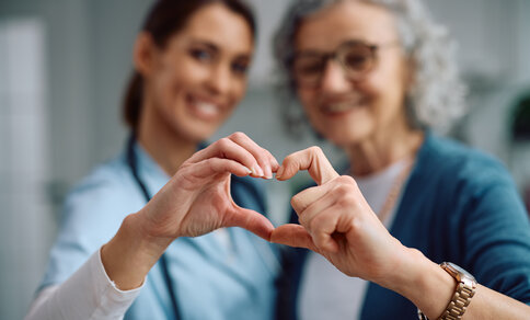 A young doctor in blue scrubs and an elderly patient with grey hair and glasses form a heart shape together with their hands in the foreground, both smiling softly out of focus in the background.