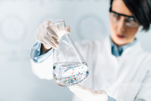 A scientist in a laboratory carefully holds a large Erlenmeyer flask filled with a clear liquid. Small colourful particles are floating in the liquid. She is wearing safety goggles, white gloves and a lab coat. The background is blurred, which draws the focus to the flask and the particles in the liquid. 