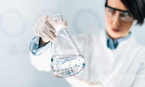 A scientist in a laboratory carefully holds a large Erlenmeyer flask filled with a clear liquid. Small colourful particles are floating in the liquid. She is wearing safety goggles, white gloves and a lab coat. The background is blurred, which draws the focus to the flask and the particles in the liquid. 