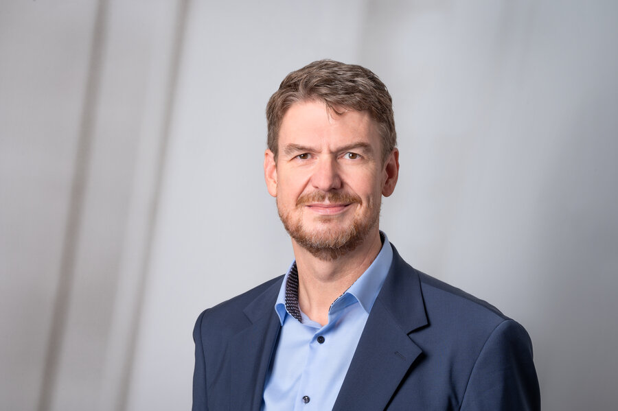 A middle-aged man in a light blue shirt and a navy blazer against a grey background.