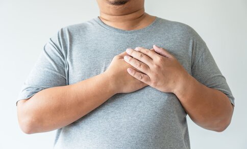 A person in a gray t-shirt places both hands over their chest, conveying concern or discomfort. 