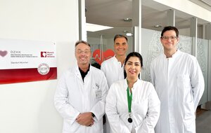 Group photo of the TRIC-I-HF study team standing in front of a DZHK (German Centre for Cardiovascular Research) sign at the Munich site, in white lab coats: three men at the back and one woman wearing a green lanyard in the front centre.