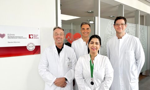 Group photo of the TRIC-I-HF study team standing in front of a DZHK (German Centre for Cardiovascular Research) sign at the Munich site, in white lab coats: three men at the back and one woman wearing a green lanyard in the front centre.