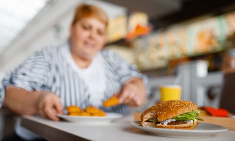 Eine stark übergewichtige Frau sitzt an einem Tisch, vor ihr ein Hamburger und eine Portion Pommes frites.