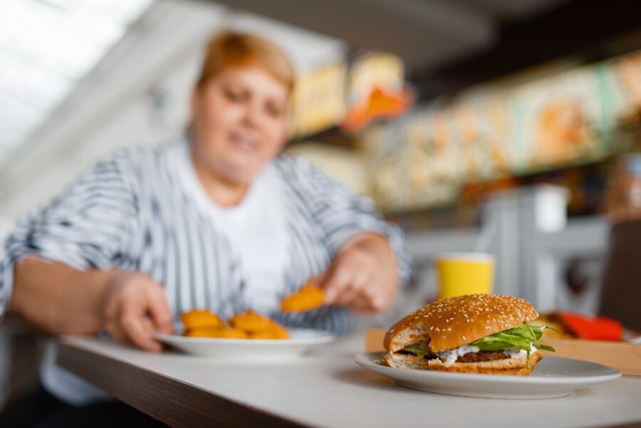 A very overweight woman sits at a table with a hamburger and a portion of chips in front of her.