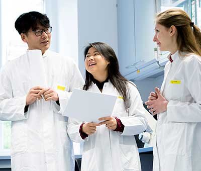 Three young professionals in white lab coats talk in a medical or scientific environment while one person holds a clipboard.’