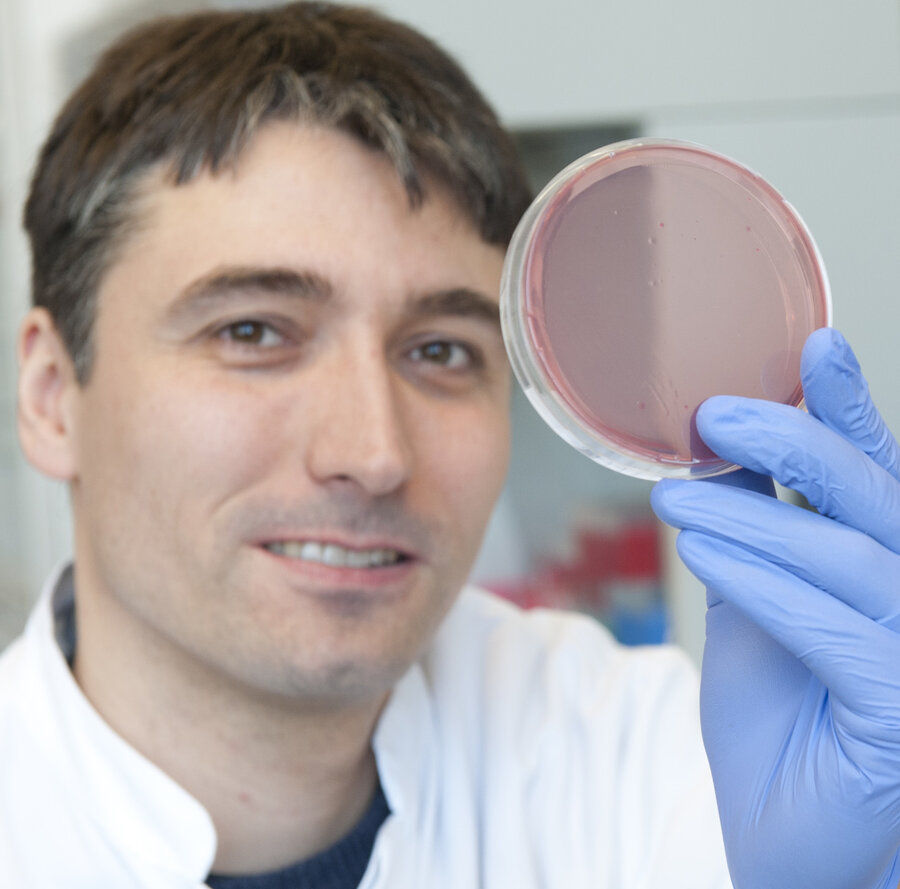 A scientist in a white lab coat, it is Prof Daniel Reinhardt, is holding a petri dish with a reddish substance in his right hand, which is covered with a blue glove. He looks into the camera and smiles slightly. The surroundings suggest a laboratory; blurred laboratory utensils can be seen in the background. 