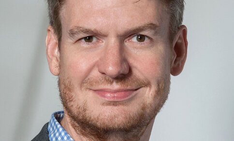 Portrait photo of a man with curly, dark blond hair and a beard, wearing a blue-and-white checked shirt and a grey blazer, smiling at the camera against a neutral light grey background.