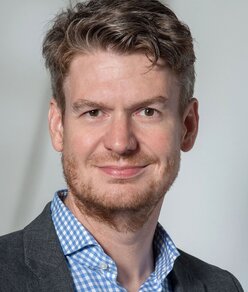 Portrait photo of a man with curly, dark blond hair and a beard, wearing a blue-and-white checked shirt and a grey blazer, smiling at the camera against a neutral light grey background.