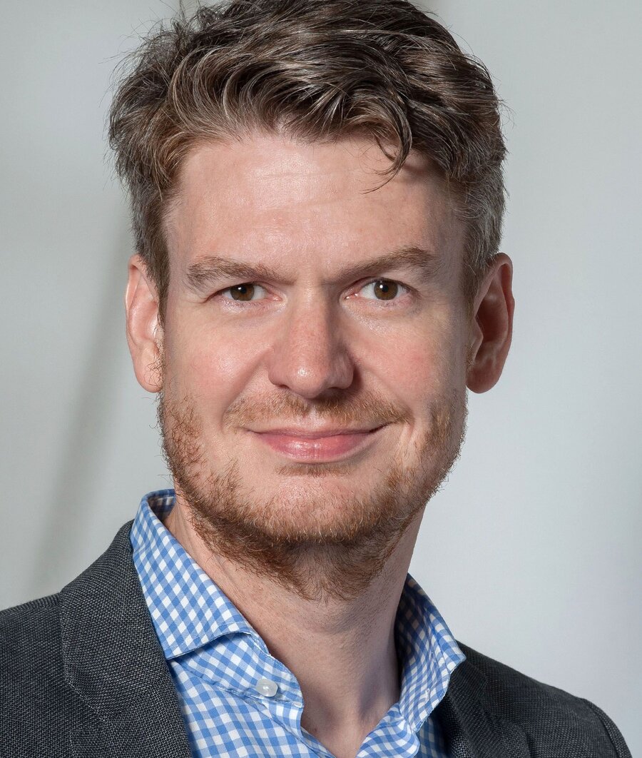 Portrait photo of a man with curly, dark blond hair and a beard, wearing a blue-and-white checked shirt and a grey blazer, smiling at the camera against a neutral light grey background.