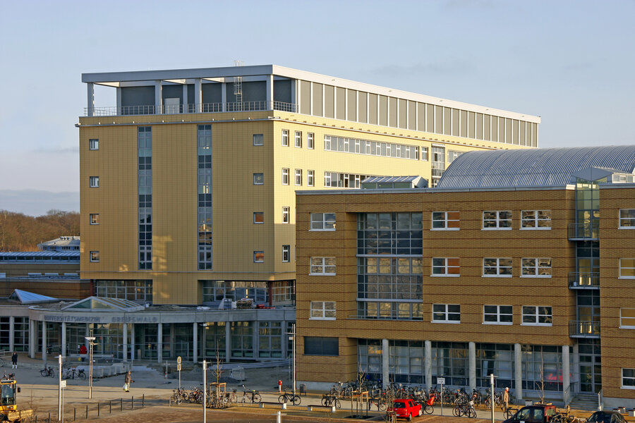 Modern academic building with yellow and brown facades, large windows, and bicycles parked outside on a sunny day.