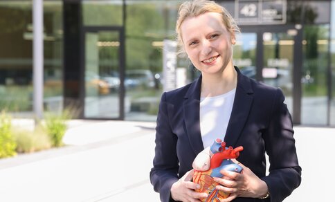 Eine junge Frau im Hosenanzug - identifziert als Prof. Dr. Constanze Schmidt - hält ein anatomisches Modell eines Herzens in der Hand und lächelt. Im Hintergrund ist der Eingangsbereich der Klinik für Kardiologie der Universitätsmedizin Göttingen zu sehen. 