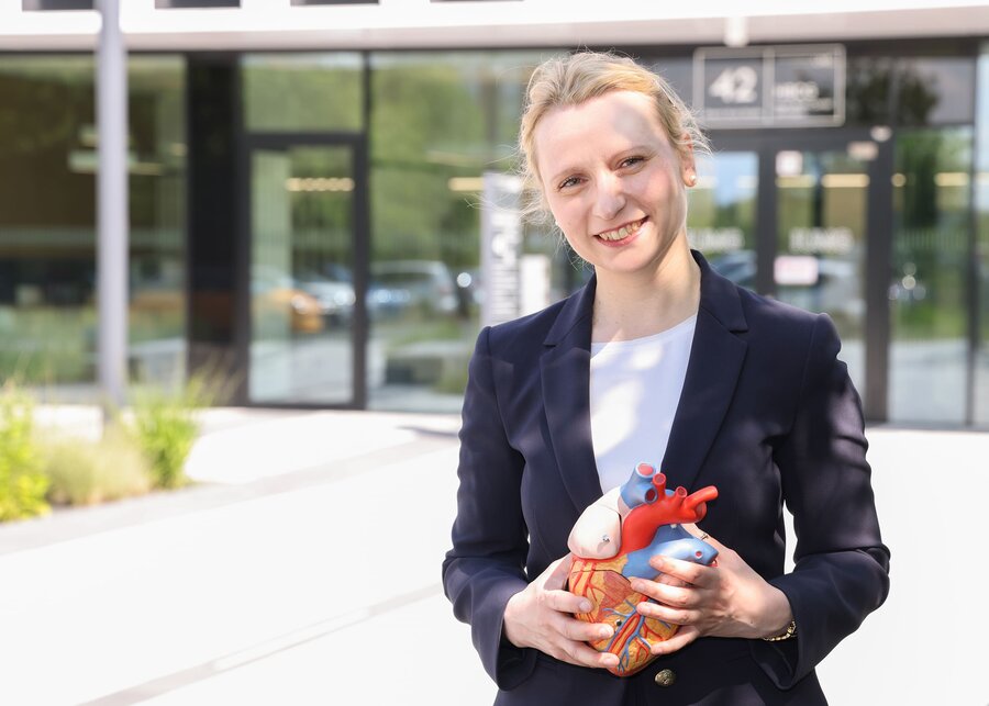 A young woman in a trouser suit - identified as Prof Dr Constanze Schmidt - holds an anatomical model of a heart in her hand and smiles. The entrance to the Cardiology Clinic at Göttingen University Medical Centre can be seen in the background.