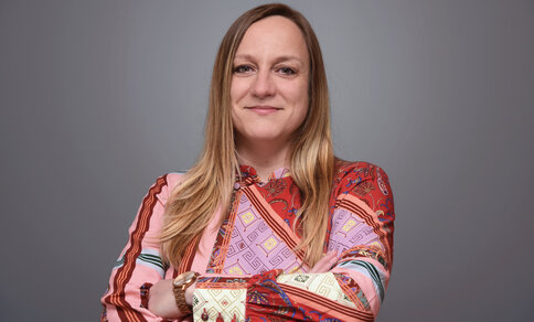  Portrait photo of Prof Daniela Panáková, smiling and looking into the camera with her arms crossed. She is wearing a colourful, patterned top and a wristwatch. Her blonde hair falls loosely over her shoulders and the background is a neutral grey. 