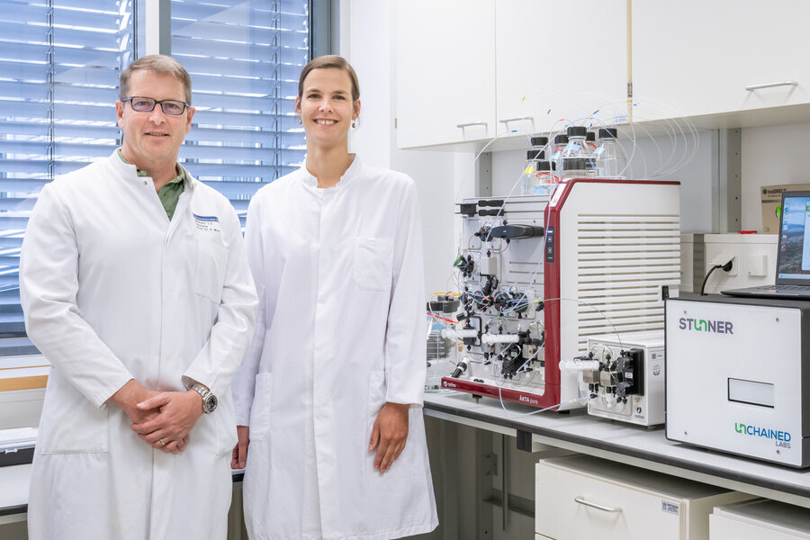 Two scientists in lab coats smile in a laboratory with advanced analytical equipment. The setting conveys a tone of professionalism and innovation.