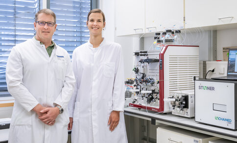 Zwei Wissenschaftler in Laborkitteln lächeln in einem Labor mit modernsten Analysegeräten. Die Umgebung vermittelt einen Eindruck von Professionalität und Innovation.
