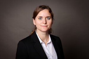 Portrait photo of researcher Jana Grune, with reddish-brown hair tied back, wearing a black blazer and white blouse against a brown background.