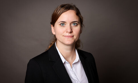 Porträtfoto der Wissenschaftlerin Jana Grune mit rot-braunen, zurückgebundenen Haaren, schwarzem Blazer und weißer Bluse vor braunem Hintergrund.