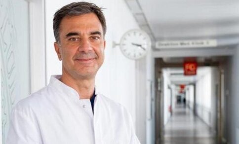 A man (Prof. Dr Steffen Massberg) is standing in a corridor wearing a lab coat and looking friendly at the camera.