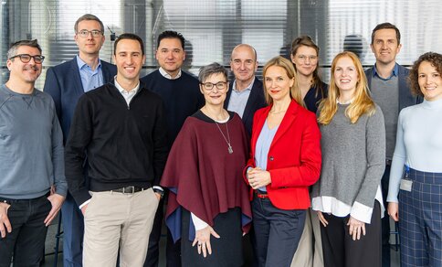 The ARC project team: Eleven people, five men and six women, stand in front of glass walls in a modern office building. They smile at the camera and appear friendly and professionally dressed. Some wear suits or blazers, others casual business attire.