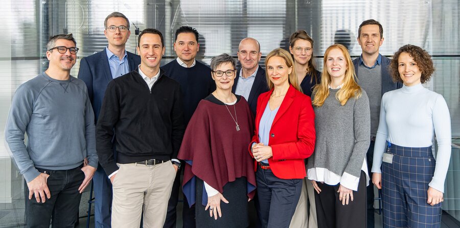The ARC project team: Eleven people, five men and six women, stand in front of glass walls in a modern office building. They smile at the camera and appear friendly and professionally dressed. Some wear suits or blazers, others casual business attire.