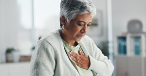 Elderly woman with short gray hair and a concerned expression clutches her chest, suggesting discomfort. 