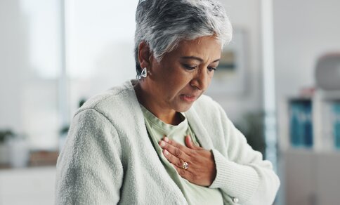 Elderly woman with short gray hair and a concerned expression clutches her chest, suggesting discomfort. 