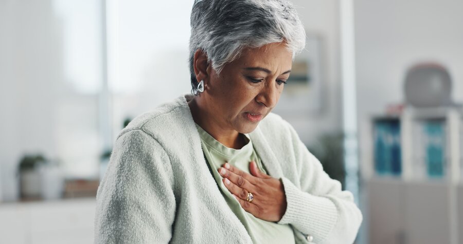 Elderly woman with short gray hair and a concerned expression clutches her chest, suggesting discomfort. 