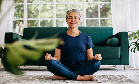  **Alt-Text:** Elderly woman sits relaxed cross-legged on a yoga mat in the living room and meditates with her eyes closed - symbolising mindfulness and mental well-being as a contribution to heart health.