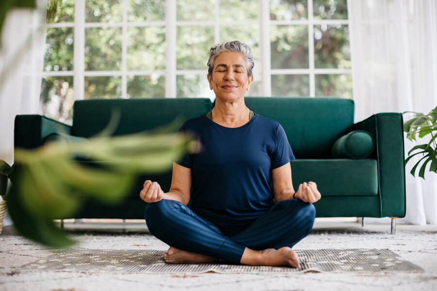 **Alt-Text:**   Ältere Frau sitzt entspannt im Schneidersitz auf einer Yogamatte im Wohnzimmer und meditiert mit geschlossenen Augen – Symbol für Achtsamkeit und psychisches Wohlbefinden als Beitrag zur Herzgesundheit.